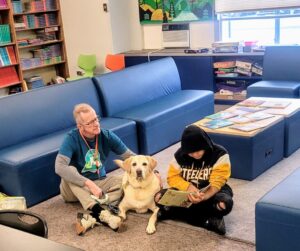 A man and a boy sit in a school library with a Labrador Retriever between them. The boy is reading a book to the dog.