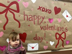 A toddler sits in front of a Valentine's Day banner with oversized heart-shaped glasses in his hand.