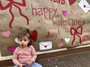 A toddler sits in front of a Valentine's Day banner.