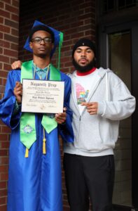Two young men stand next to each other, one wearing a cap and gown and holding a Nazareth Prep diploma.