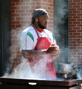 A young man stands outside at a grill with steam rising around him